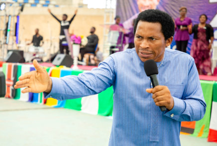 a man in a blue shirt, holding a microphone and speaking to a crowd